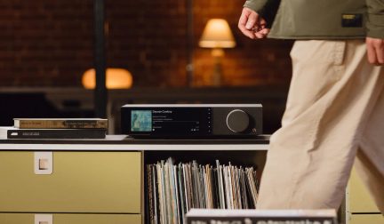 Lettore audio di rete nero con grande schermo a colori e manopola centrale appoggiato su un mobile per vinili in un soggiorno
