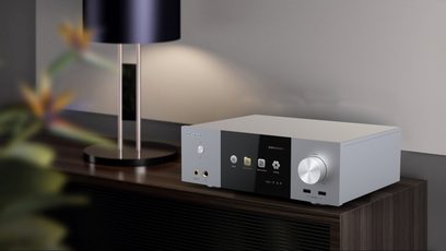 Sølvfarvet Zidoo Neo S multimedie-afspiller, set forfra, placeret på et mørkebrun træmøbel, med en sort lampe i baggrunden
