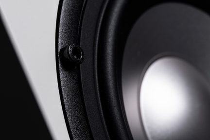 Zväčšený šikmý záběr čierneho reproduktora z kovu a plastu, upevňovacie skrutky na textúrovanom ráme, lesklý kruhový kužeľ