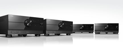 Four black amplifiers aligned in a diagonal perspective with circular knobs and digital screens on a reflective surface
