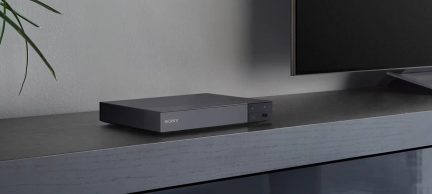 Sort, rektangulær medieafspiller på gråt møbel, Sony-logo synligt, sort skærm til højre, grøn plante til venstre