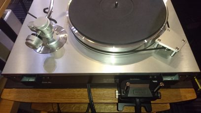 Turntable with tonearm and cartridge mounted on a brushed metal platter, placed on a light wood cabinet