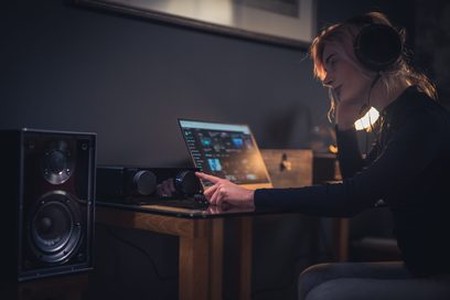 Kvinde siddende foran en åben bærbar computer, der bruger en lydmixerkonsol i et mørkt optagelsesstudie
