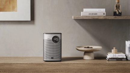 Harmaa sylinterimäinen projektori puupöydällä, seinähylly kirjoilla, puuveistos, kivivati käpyjen kanssa