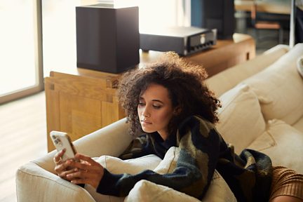 Person med krøllet hår liggende på beige stofsofa, sort og grøn sweater, gylden smartphone i hånden, 3/4-visning