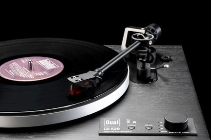 Giradischi nero con finitura effetto legno visto da tre quarti frontale, piatto metallico argentato con disco nero, braccio di lettura tubolare nero con testina rossa traslucida, comandi e ampia manopola neri sul frontale