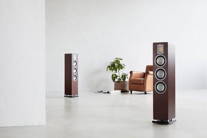 Sloupová reprosoustava AudioVector QR5 ořech, klubové křeslo a zelená rostlina, na bílém podkladu, pohled zepředu