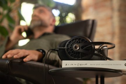 Czarny kask na książce "Room to Dream" na stole, rozmyty mężczyzna w tle, fotel, rośliny, ściana z cegieł