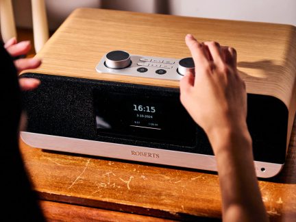 Roberts clockradio i lyst træ med digitalt display på fronten og store metal-knapper, hånd der justerer den øverste knap