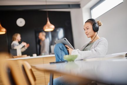 Persona seduta con cuffie audio, tenendo un tablet, due persone sfocate sullo sfondo, vista laterale