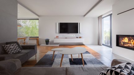 Forside af en moderne stue med et gråt hjørnesofa i stof og puder med mønstre, et sofabord med hvide bordplader på lyse træben, et hvidt lakket tv-møbel med stort sort skærm indrammet af to hvide højttalere og en lydbar, et glasdørsbrændeovn til højre, trægulv delvist dækket af et geometrisk gråt og sort tæppe