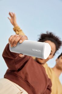 Bílý bezdrátový reproduktor Sonos Roam SL držený v ruce na světle modrém pozadí