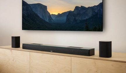 Zwarte Canton-soundbar met twee compacte luidsprekers op een licht houten meubel onder een wandtelevisie met een berglandschap op het scherm
