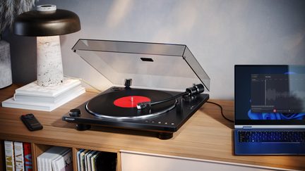 Black vinyl turntable with metal and plastic construction, transparent lid raised, black record with red label, three-quarter view