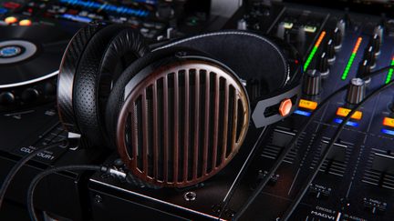 Over-ear hovedtelefoner i brunt træ med åbne skaller med lodrette gitter, sorte læderørepuder og sort metalbøjle, set i tæt nærbillede fra trekvart forfra placeret på et mixerbord