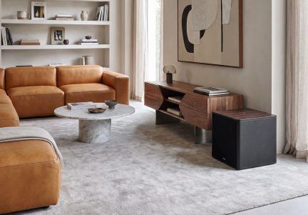 Kompaktní černý subwoofer s hnědým vrškem, prezentovaný v moderním obývacím pokoji s karamelovou pohovkou a TV stolkem