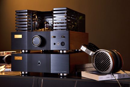 Tredjehåndsvisning av en hi-fi rørforsterker i to stablede enheter, frontpanel i børstet svart aluminium med stor gullinnrammet dreieknapp og hodetelefonutganger, brune tresider toppet med et sort perforert metallgitter, ledsaget til høyre av et sølvfarget, åpent circum-auralt hodesett med svarte puter som hviler på bøker