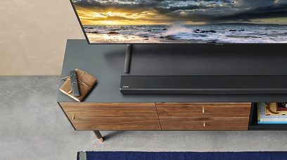 Træ-TV-møbel med skuffer, sort Samsung HW-R530 soundbar, fjernbetjening, ovenfra set, TV-skærm der viser en solnedgang over havet
