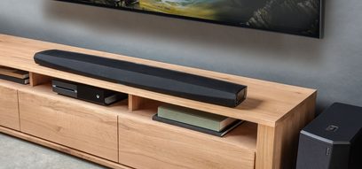 Czarna, prostokątna, wydłużona soundbar Denon DHT-S716H z widocznymi głośnikami, umieszczona na jasnej drewnianej szafce pod telewizorem