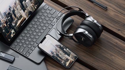 Kulak üstü siyah ve gümüş metal fırçalı ve suni deri kulaklık, üstten görünüm, ahşap bir masanın üzerinde bir akıllı telefon ve klavyenin yanında duruyor