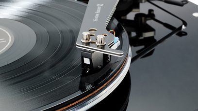 Close-up in lichte vogelvlucht op de hi-fi draaitafel Vertere Acoustics Pack DGX Dark Sabre, met een glanzend zwart plateau omrand door acryl, een opengewerkte metalen arm met twee chromen schroeven, een zwarte cartridge en duidelijk zichtbare groeven op de plaat.
