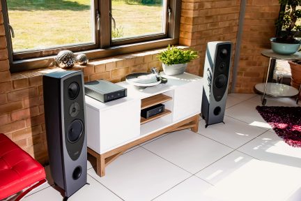 Deux enceintes noires, un meuble blanc avec platine vinyle et rangements, dans une pièce avec fenêtres et mur en briques