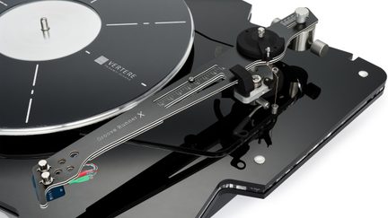 Sort, blank akryl vinylpladespiller set fra øvre trekvart vinkel, sort cirkulær pladetallerken med hvide markeringer, gråmetallic tonearm med påskriften "Groove Runner X", gradueret markør og synlige flerfarvede kabler
