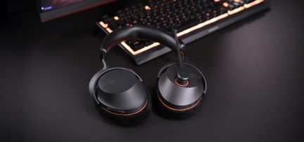 Căști de gaming negre cu microfon și accente portocalii, așezate pe birou în fața unei tastaturi mecanice iluminate din spate