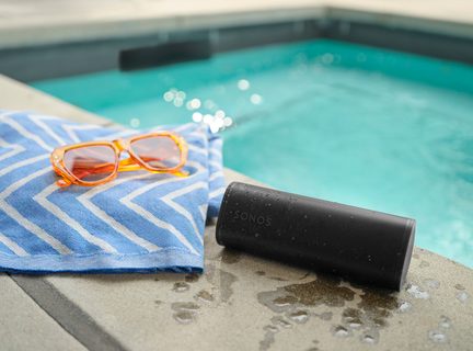 Altoparlante nero, asciugamano blu a motivi bianchi, occhiali da sole arancioni, bordo piscina