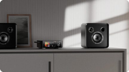 Black hi-fi system with a color display and two speakers with rounded corners, placed on a gray sideboard