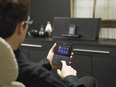 Ruka držící černý dálkový ovladač Sony s dotykovým displejem a modrým uživatelským rozhraním, v tmavém interiéru