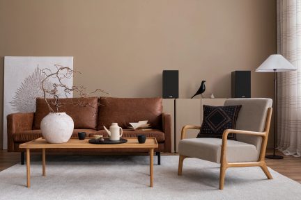 Salotto con divano in pelle marrone, poltrona in legno e tessuto beige, tavolino in legno, vaso bianco, altoparlanti neri, lampada da terra bianca