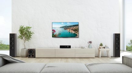 Living cu perete alb texturat, televizor montat pe perete, mobilier jos bej, 2 boxe negre, subwoofer, plante, măsuță de cafea