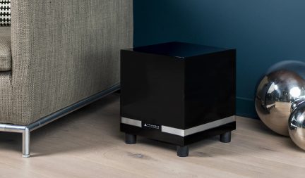 Cassa subwoofer cubica in legno con finitura laccata nera, dotata di una fascia metallica argentata sulla parte inferiore e di quattro piedini cilindrici, vista in prospettiva frontale destra