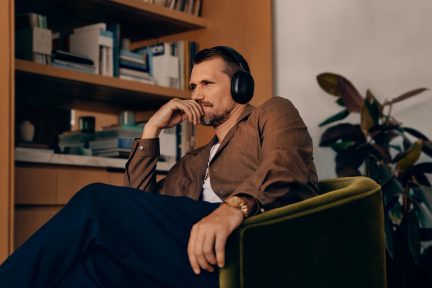 Vīrietis, sēžas zalā krēslā, ar brūna kājas un zilu pukstu, uz galvas nāk audio ūdens katra melns, ar zelta pulksteni un grāmatu skapiem pašreizējā fona