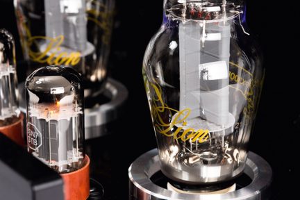 Primo piano obliquo di due tubi elettronici cilindrici a forma di bottiglia, con involucro in vetro trasparente dai riflessi argentati, elementi interni metallici, iscrizione dorata « Lion » sul più grande, base circolare argentata e collarino marrone sul più piccolo da cui emana una leggera luce arancione