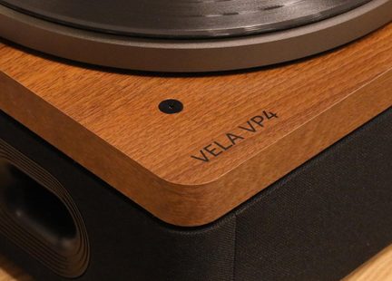 Detail gramofonové desky z hnědého dřeva, šedý kovový talíř, černý šroub a označení „VELA VP4“, záběr v detailním šikmém pohledu