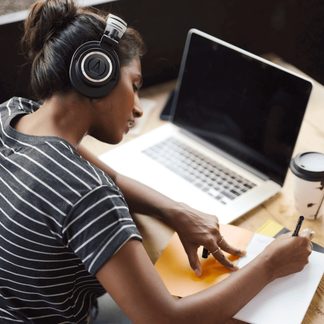 Osoba s černými sluchátky, píšící na oranžovém a bílém papíře, notebook a hrnek na dřevěném stole