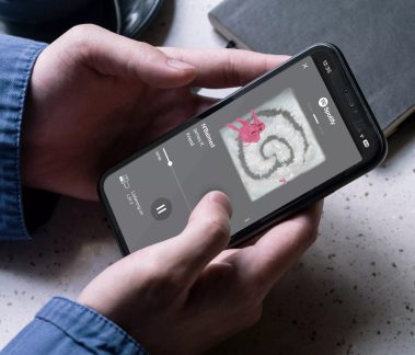Zwarte smartphone met Spotify-app, met twee handen vastgehouden boven een lichte tafel, blauwe overhemdsmouwen zichtbaar bij de polsen