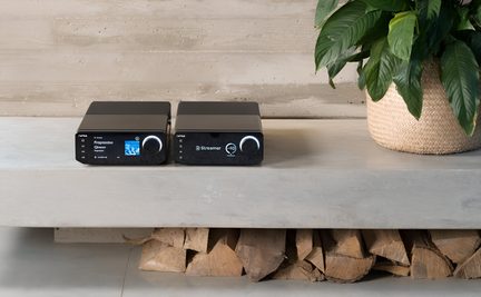 Dois módulos de áudio pretos em metal e plástico, com ecrãs e botões, vistos de frente, sobre uma prateleira de betão junto a uma planta
