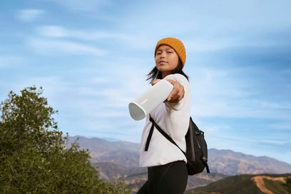 The image shows a young Asian woman holding a white portable speaker in a sunny mountainous landscape, wearing a yellow beanie and a black backpack