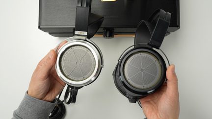 Két fekete és ezüst nyitott Stax fejhallgató kézben tartva egy fekete fejhallgató-erősítő előtt