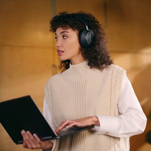 Ung kvinde iført en hvid sweater, der lytter til musik med et sort over-øre hovedsæt, arbejdende på en bærbar computer