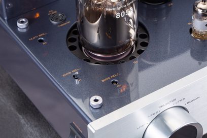 Detailní záběr mírně shora na černou elektronkovou zesilovač Cayin Jazz 100 s černým lesklým kovovým šasi a stříbrnou kartáčovanou hliníkovou přední stranou, který zdůrazňuje skleněnou elektronku 805A obklopenou kruhovými perforacemi, přepínač pro nastavení biasu a velký válcový ovládací knoflík hlasitosti.