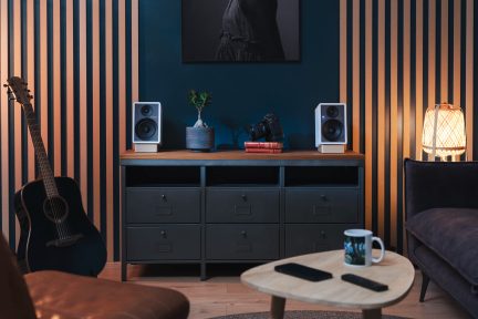 Zwarte staande akoestische gitaar, grijze lage kast met 6 lades, 2 witte luidsprekers, gevlochten lamp, licht houten salontafel