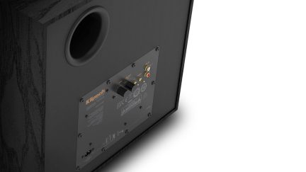 Sort højtaler med træfinér, bagside set fra venstre skråt bagfra, med cirkulær øvre ventilationsåbning og metalplade med to drejeknapper samt røde og hvide RCA-stik