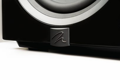 Dynamo 12 subwoofer i sort lak, delvis set fra en lav vinkel, skinnende sort overflade, metallisk logo i relief, synlig cirkulær højttalerkant