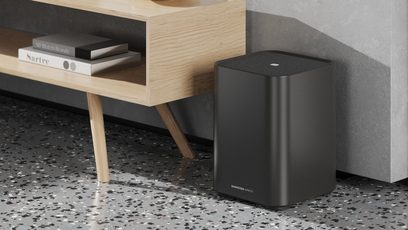Sennheiser AMBEO Sub mat siyah dikdörtgen subwoofer, açık renk ahşap bir mobilyanın yanında yerde, ön görünüm