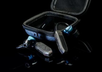 Trekvartersvisning af en åben, grå-antracit stofetui med lynlås, der indeholder to trådløse sorte plastik-ørestykker med metallisk blå skiver og et sort rektangulært modul med hvide lysdioder