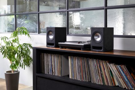 Dos altavoces negros, tocadiscos, estante con discos, planta verde en maceta, ventanas de vidrio texturizado
